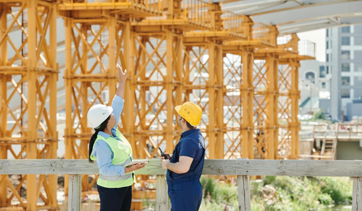 Two workers, one female and one male, stand in front of large yellow scaffolding. The woman wears a bright yellow safety vest and a white hat. She is pointing to something up high and carrying a notepad. The man wears a dark blue jumpsuit and yellow hard hat and is carrying a walkie talkie.